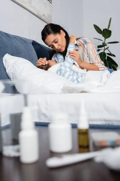 Criança doente abraçando mãe enquanto deitado na cama perto da mesa com medicamentos em primeiro plano desfocado — Fotografia de Stock