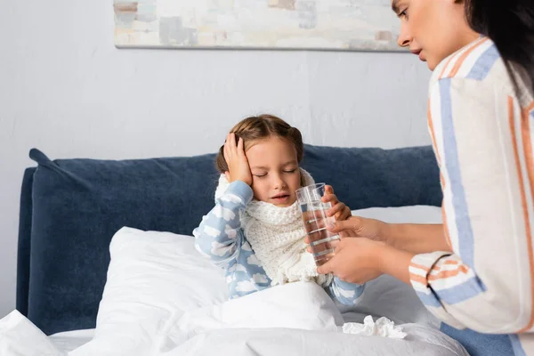 Madre dando vaso de agua al niño enfermo que sufre de dolor de cabeza mientras está sentado en la cama con los ojos cerrados - foto de stock