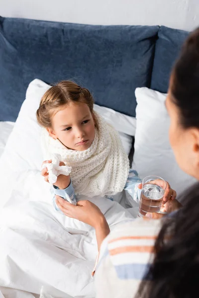 Mulher dando vidro de água para a filha doente segurando guardanapo de papel, foreground borrado — Fotografia de Stock