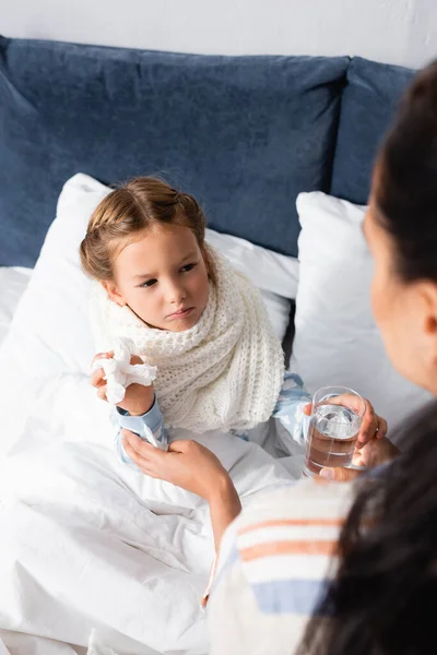 Donna che dona un bicchiere d'acqua alla figlia malata con in mano un tovagliolo di carta, primo piano sfocato — Foto stock