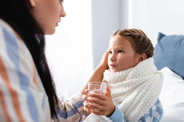 Sick girl holding glass of water and looking at mother on blurred foreground — Stock Photo