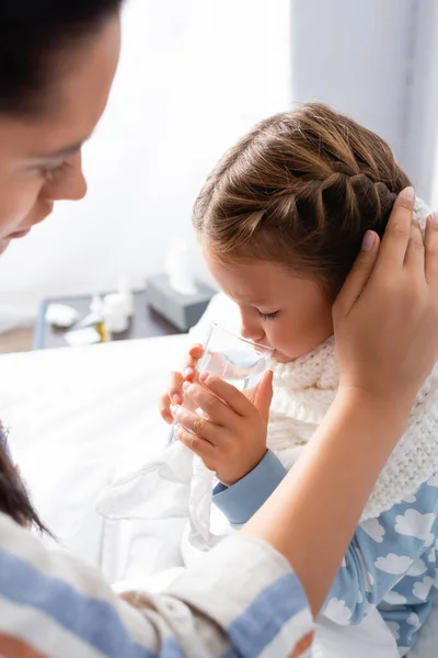 Krankes Kind trinkt Wasser, während Mutter ihren Kopf auf verschwommenem Vordergrund berührt — Stockfoto