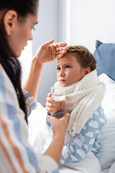 Woman giving glass of water to ill daughter while touching her forehead on blurred foreground — Stock Photo