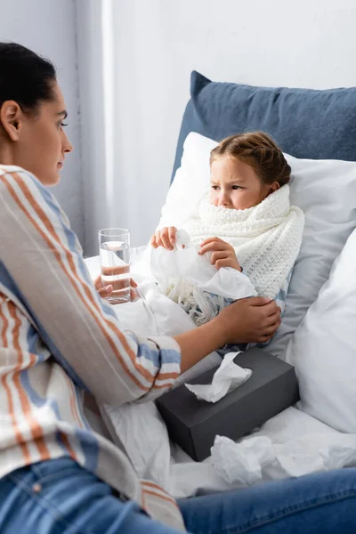 Woman holding glass of water while sitting near upset, diseased daughter — Stock Photo