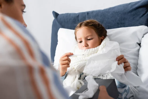 Fille malade regardant thermomètre près de mère tenant des serviettes en papier sur le premier plan flou — Photo de stock