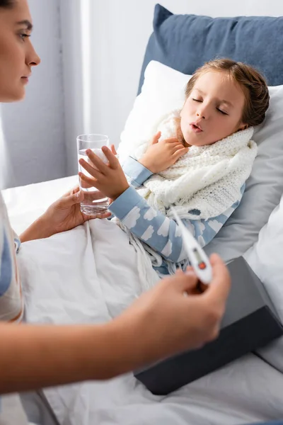 Madre sosteniendo el termómetro y dando agua a la niña enferma acostada en la cama con los ojos cerrados, borrosa primer plano - foto de stock