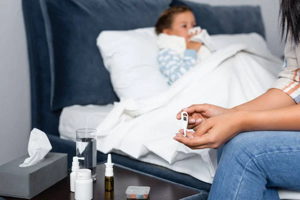 Woman looking at thermometer near sick child lying in bed on blurred background — Stock Photo