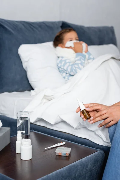 Woman holding nasal spray near bedside table with medicines and sick daughter lying in bed on blurred background — Stock Photo