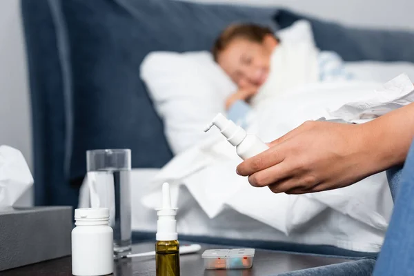 Woman holding throat spray near bedside table with medicines and sick daughter lying in bed on blurred background — Stock Photo