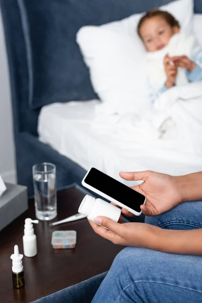 Mujer sosteniendo teléfono inteligente y botella de pastillas cerca de hija enferma acostada en la cama sobre fondo borroso - foto de stock