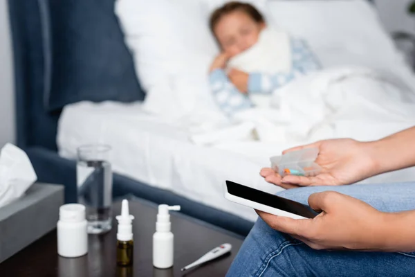 Mujer sosteniendo teléfono inteligente y pastillas mientras está sentado cerca de un niño enfermo acostado en la cama sobre un fondo borroso - foto de stock