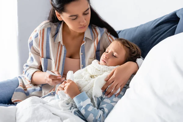 Женщина держит термометр и обнимает больную дочь лежащую в постели с закрытыми глазами — стоковое фото