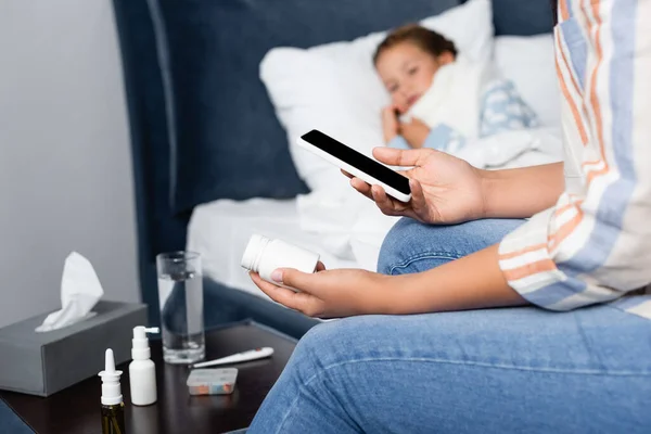 Mãe segurando garrafa de pílulas e telefone celular perto da filha doente deitada na cama — Fotografia de Stock