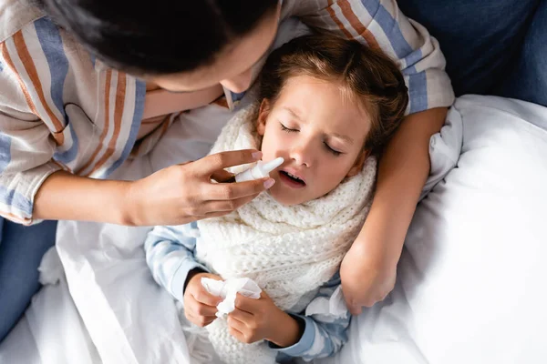 Visão superior da criança doente deitada com os olhos fechados enquanto a mãe usa spray nasal — Fotografia de Stock
