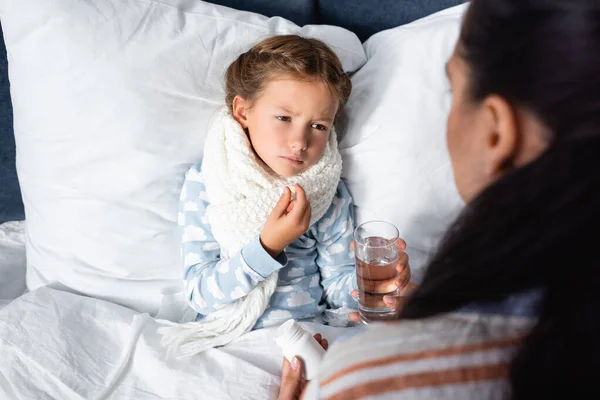 Sick girl holding pill near mother holding glass of water on blurred foreground — Stock Photo
