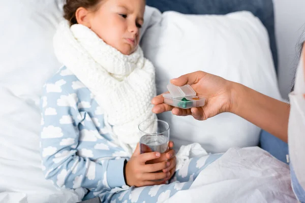 Mother giving pills to sad, diseased daughter holding glass of water on blurred background — Stock Photo