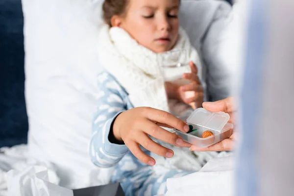 Woman giving medicines to daughter lying in bed and holding glass of water on blurred background — Stock Photo
