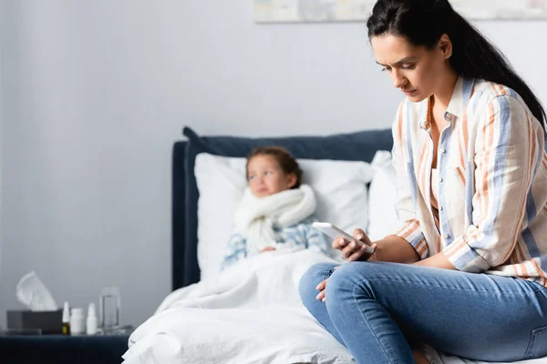 Upset woman calling by smartphone while sitting on bed near ill daughter on blurred background — Stock Photo