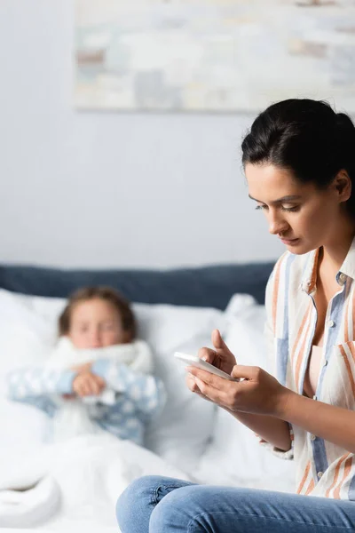 Сообщения матери на мобильном телефоне рядом с больной дочерью, лежащей в постели на размытом фоне — стоковое фото