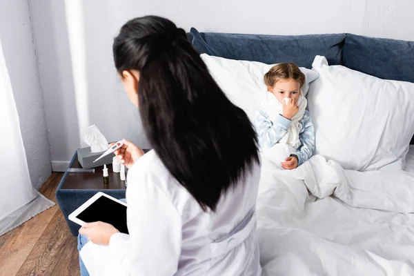 Médico mirando el termómetro cerca de niña enferma acostada en la cama - foto de stock