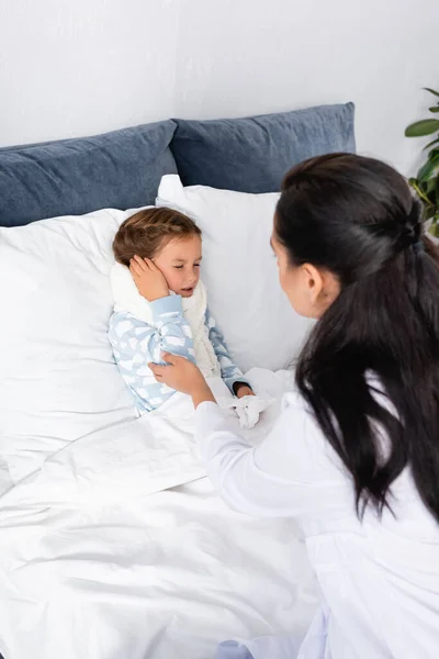 Médico tocando niño enfermo que sufre de dolor en el oído mientras está acostado en la cama - foto de stock