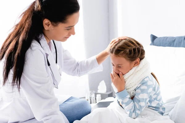 Doctor touching head of squeezing girl sitting in bed — Stock Photo