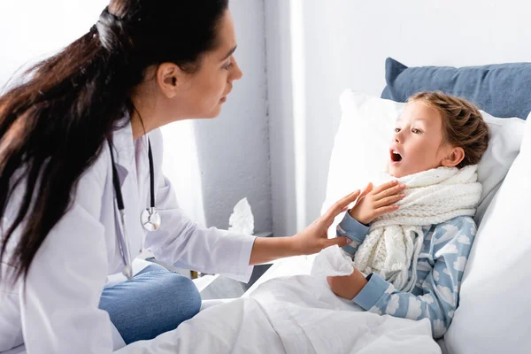 Pediatrician examining sore throat of diseased girl lying in bed — Stock Photo