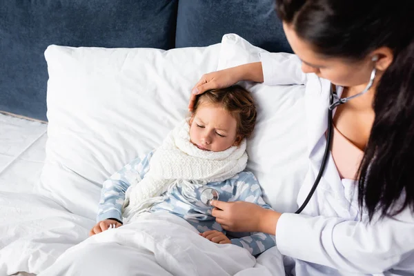 Vue grand angle du pédiatre touchant la tête de la fille malade et l'examinant avec stéthoscope sur le premier plan flou — Photo de stock