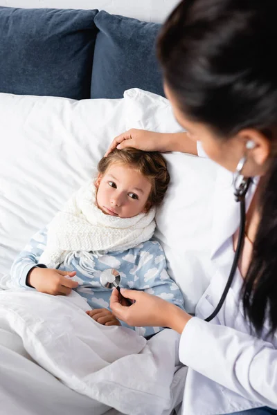 Arzt berührt krankes Mädchen am Kopf, liegt im Bett und untersucht sie mit Stethoskop — Stockfoto