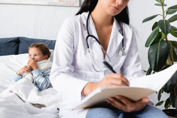 Diseased child wiping nose near pediatrician writing in outpatient card on blurred foreground — Stock Photo