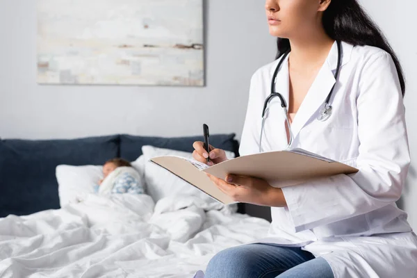 Pediatrician writing in outpatient card near ill girl lying in bed — Stock Photo