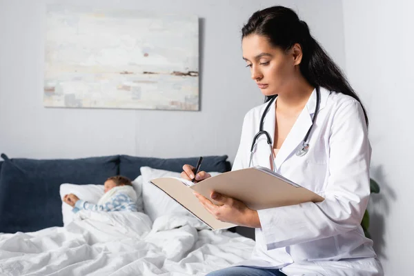 Médico serio escribir diagnóstico cerca de niño enfermo acostado en la cama - foto de stock