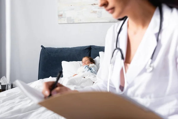 Ill child lying in bed while pediatrician writing diagnosis on blurred foreground — Stock Photo