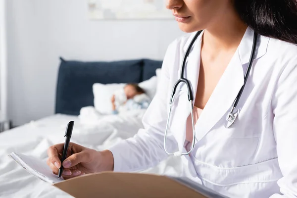 Pediatra escribiendo diagnóstico mientras niña enferma durmiendo en la cama sobre fondo borroso - foto de stock
