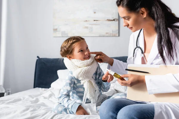 Pediatrician holding nasal spray while touching nose of smiling girl in warm scarf — Stock Photo