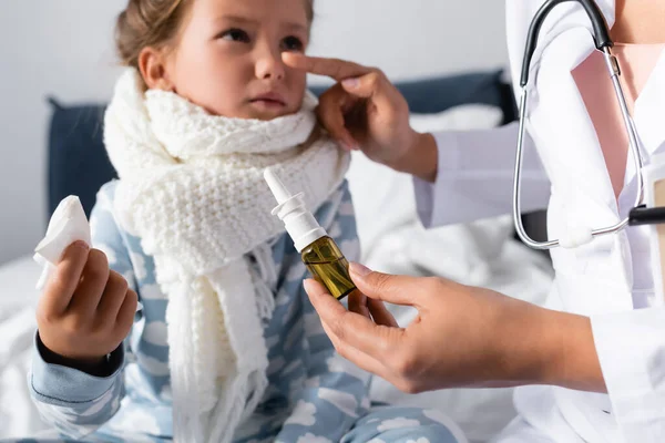Doctor pointing at nose of diseased girl while holding nasal spray on blurred background — Stock Photo