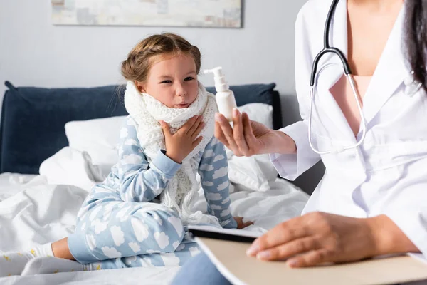 Doctor showing throat spray to displeased, sick girl in warm scarf around neck, blurred foreground — Stock Photo