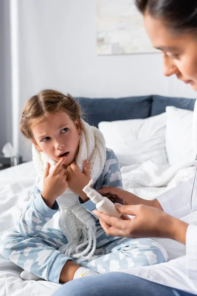 Krankes Mädchen zeigt auf offenen Mund, während Arzt Halsspray hält — Stockfoto