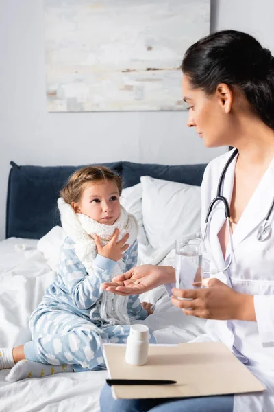 Kinderarzt gibt Medizin und Glas Wasser an krankes Mädchen, das Halsschmerzen auf verschwommenem Vordergrund berührt — Stockfoto