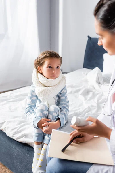 Fille malade en écharpe chaude regardant médecin tenant carte ambulatoire et bouteille avec des pilules — Photo de stock