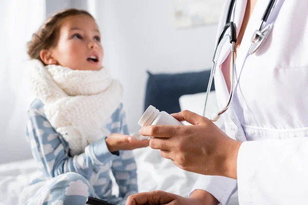 Kinderarzt gibt kranken Mädchen mit warmem Schal am Hals vor verschwommenem Hintergrund Pillen — Stockfoto