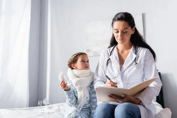 Pediatra escribiendo diagnóstico mientras niña enferma sosteniendo servilleta de papel - foto de stock