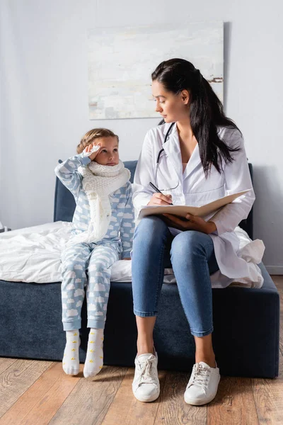 Doctor escribiendo en tarjeta ambulatoria mientras está sentado cerca de niña enferma usando pijama - foto de stock