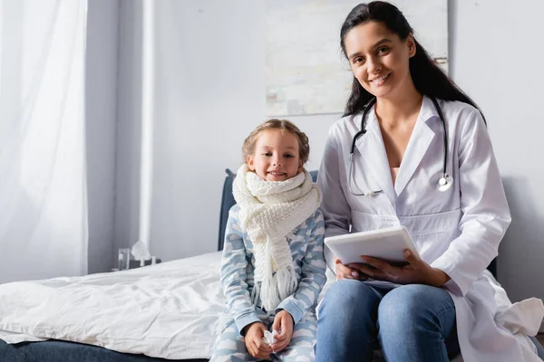 Glückliches Kind und Kinderarzt auf Bett sitzend und in die Kamera blickend — Stockfoto