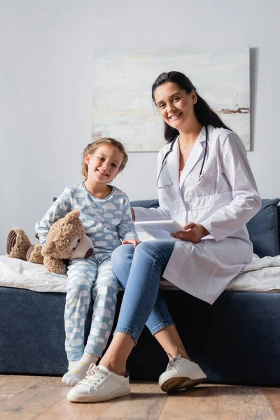 Cheerful pediatrician and child with teddy bear smiling at camera while sitting on bed — Stock Photo