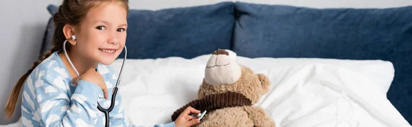 Cheerful girl looking at camera while examining teddy bear with stethoscope, banner — Stock Photo