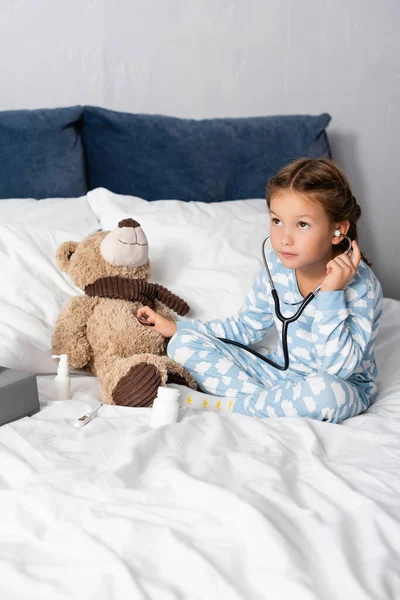 Criança brincando enquanto examina ursinho de pelúcia com estetoscópio enquanto joga na cama — Fotografia de Stock