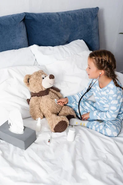 Девушка рассматривает плюшевого мишку со стетоскопом во время игры в постели — стоковое фото