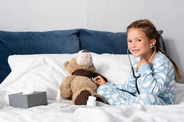 Enfant joyeux regardant la caméra tout en examinant ours en peluche avec stéthoscope — Photo de stock