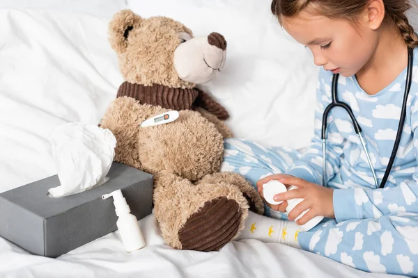 Girl opening pills bottle while playing with teddy bear in bedroom — Stock Photo